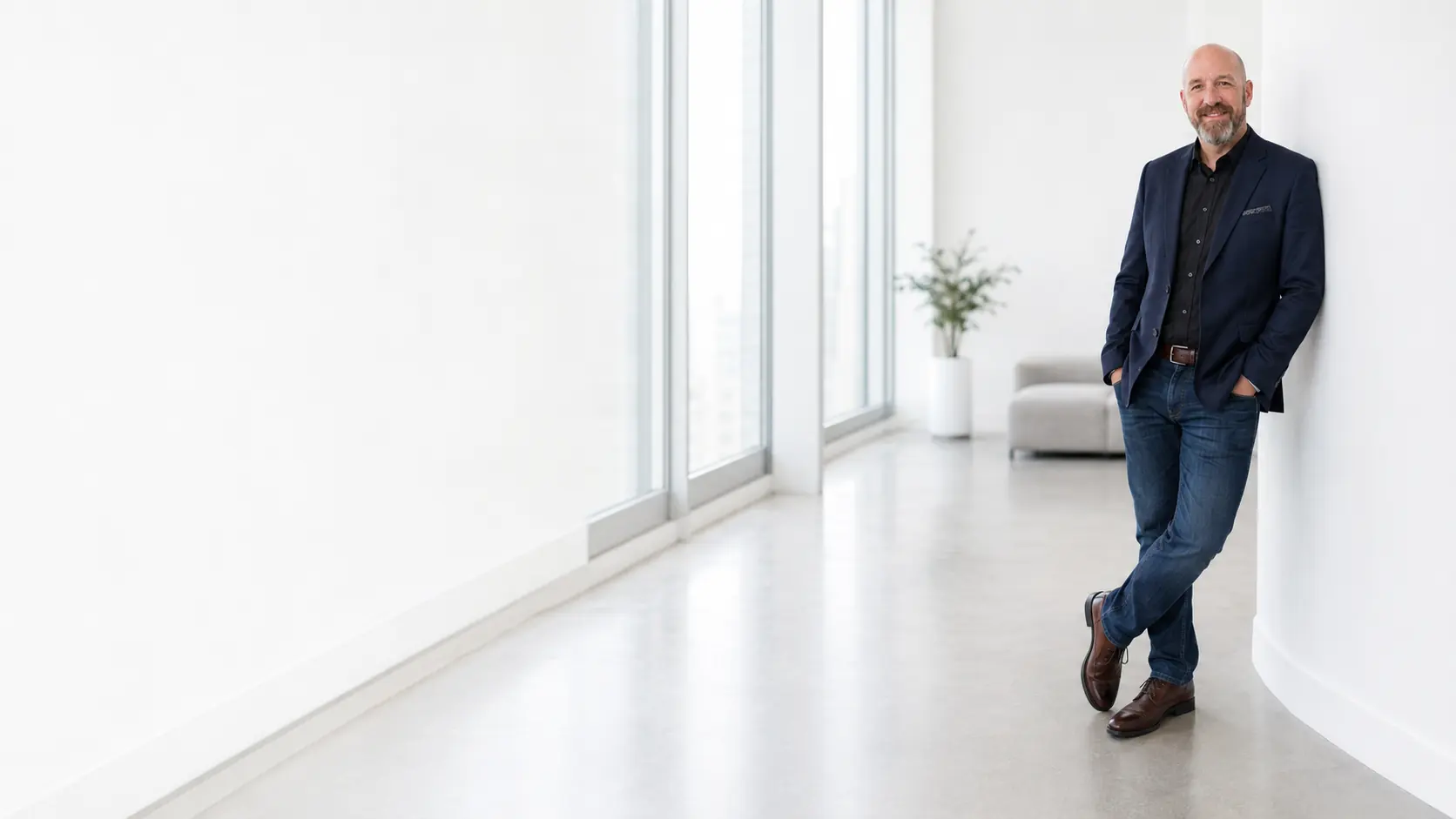 Brett Gibson standing in a bright modern office with floor-to-ceiling windows, wearing a navy jacket over a black shirt.
